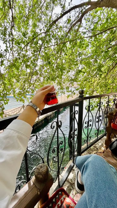 A first-person view of a hand holding a glass of red Turkish tea overlooking a waterfront through an ornate black iron railing, with another tea glass resting on a wooden chair to the side.