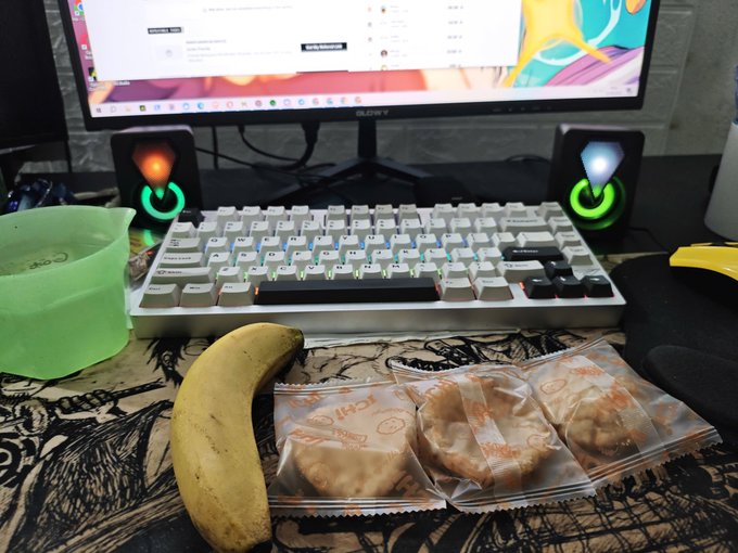 A computer desk setup with a keyboard, monitor, and RGB speakers, with a banana and three packaged pastries sitting on a patterned desk mat.