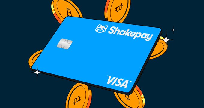 A blue Shakepay Visa credit card is shown surrounded by floating Bitcoin tokens on a dark background. The image visually represents the announcement of Shakepay's physical Visa card launch, with the Bitcoin coins emphasizing the company's cryptocurrency focus.