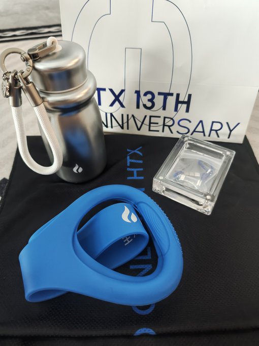 A silver insulated water bottle with carabiner, a blue silicone swim cap, and a small clear container are displayed on a black "HTX" branded pouch, with a "HTX 13TH ANNIVERSARY" card in the background, showcasing a practical sports travel gift set celebrating Huobi's 13th anniversary.