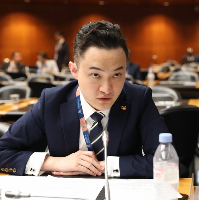 Justin Sun sits at a conference table in a dark suit and striped tie, with a small flag pin on his lapel, appearing focused as he looks toward the camera. A bottle of water and papers are visible on the table in front of him, while other attendees can be seen in the blurred background of what appears to be a formal meeting or summit.