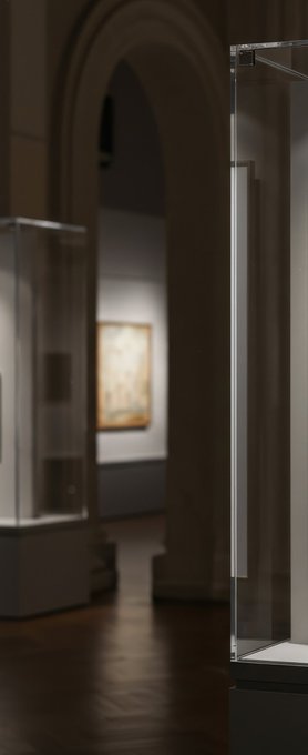 A dimly lit museum gallery featuring arched architecture, with illuminated glass display cases in the foreground and a framed artwork visible on the wall in the background.