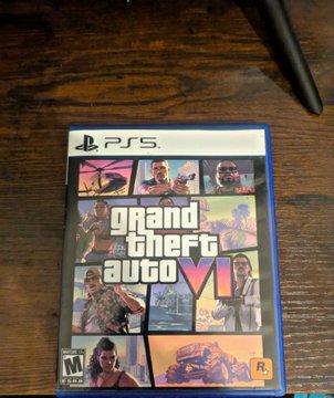 A photograph of the PS5 video game 'Grand Theft Auto VI' case resting on a wooden surface, showing the cover artwork featuring multiple characters.