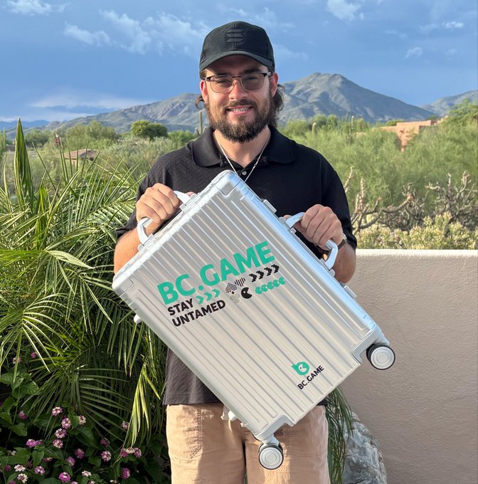 A man wearing a black cap, glasses, and BC.Game t-shirt smiling while holding a silver BC.GAME aluminum suitcase that reads 'STAY UNTAMED' outdoors with mountains and desert vegetation in the background.