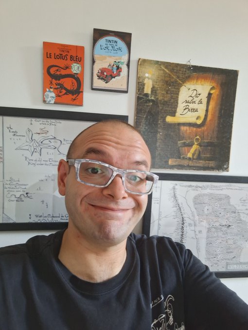 A smiling, bald man wearing white-framed glasses sits in front of a wall decorated with Tintin comic book covers ('Le Lotus Bleu' and 'L'Île Noire') and framed maps.
