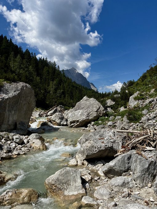 A scenic landscape photo of a rushing river flowing through large gray rocks, bordered by a forested mountain and a blue sky with white clouds.