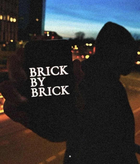A gloved hand holds up a phone with the text "BRICK BY BRICK" displayed on screen against a dusk cityscape backdrop, symbolizing the steady, patient accumulation required for long-term investment gains rather than chasing quick 100x returns.