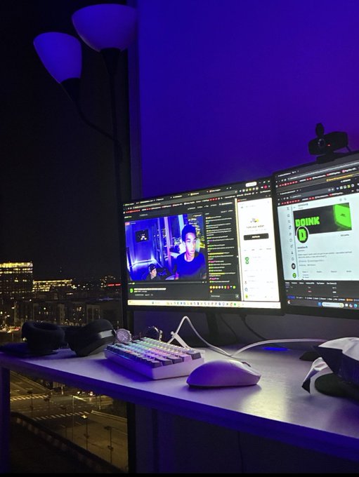 A dark gaming or streaming setup featuring dual monitors displaying a live stream with a YouTube chat on the right, illuminated by purple ambient lighting with a mechanical keyboard and mouse on the desk.