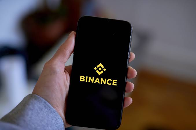 A hand holds a smartphone displaying the Binance logo (a yellow diamond made of squares) and the word 'BINANCE' against a blurred indoor background.