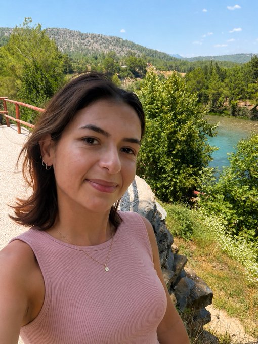 A selfie of a smiling woman in a pink tank top with shoulder-length brown hair, taken outdoors against a scenic background of turquoise water and forested mountains.