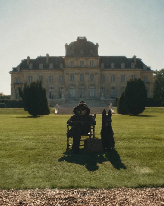 A bear wearing a hat sits in a chair on the manicured lawn of a grand European château, accompanied by a black German Shepherd dog and a briefcase, evoking a whimsically sophisticated, almost surreal aristocratic "vibe at 4B." The stately mansion backdrop and formal garden amplify the pretentious yet playful mood referenced in the caption.