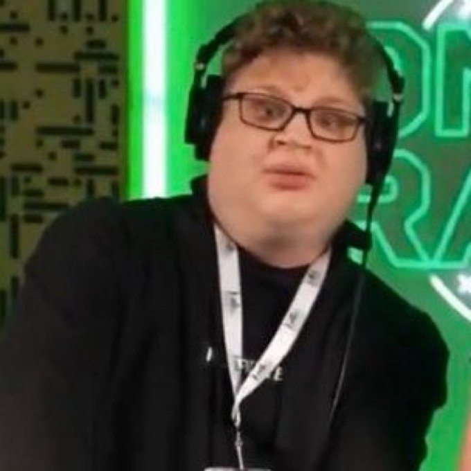 A young man with glasses and large black headphones sits in front of a green-lit background with circuit board patterns, wearing a black shirt with a lanyard.