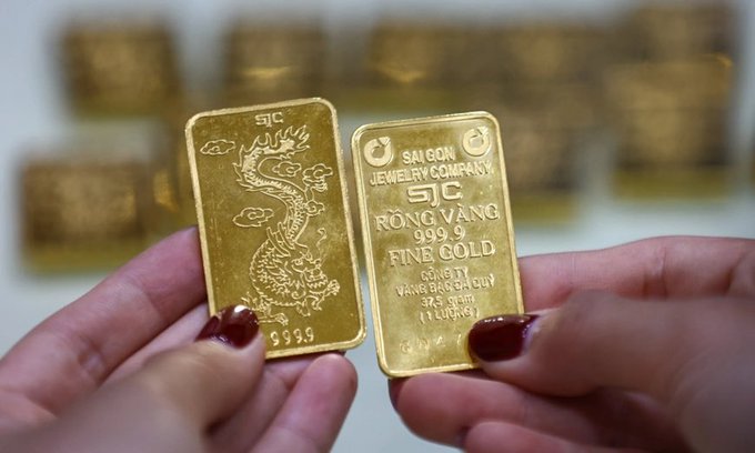 Two hands holding up gold bars from SJC (Saigon Jewelry Company), with one displaying a dragon design marked 999.9 and the other labeled 'RONG VANG FINE GOLD 999.9,' with stacks of more gold bars blurred in the background.