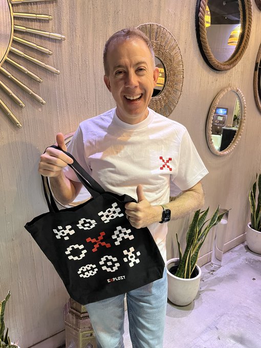 A man in a white t-shirt smiling while holding up a black tote bag with pixel-art text reading 'CODE IS POETRY' and a red X logo, standing in a room with decorative mirrors and plants.