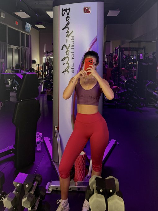 A woman takes a mirror selfie in a gym, wearing a pink sports bra and red leggings, while holding her phone in front of a poster that reads "Bring yo' a** to hip" and "RMR 2023." The "gm" post reflects a casual, morning gym vibe, emphasizing fitness motivation and starting the day strong.