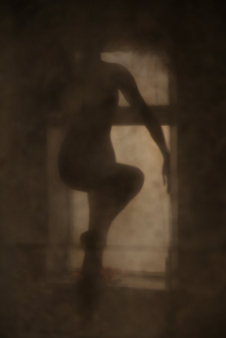 A moody, silhouetted artistic rendering of a figure posing in front of a dimly lit, arched window.