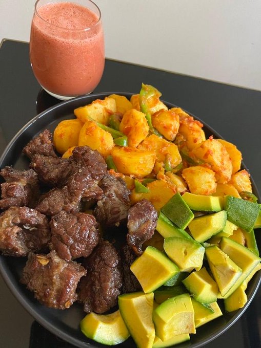 A black plate filled with chunks of seared beef, seasoned yellow potatoes with green peppers, and sliced avocado, served alongside a glass of pink fruit smoothie on a dark countertop.