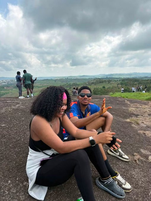 A couple sits on a rocky outcrop overlooking a vast landscape, with other people visible in the background.