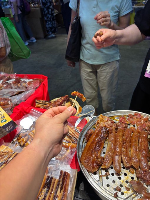A hand holds out a sample of dried meat on a toothpick at a street market stall, where smoked sausages and cured meats are displayed on a red-clothed table and a metal tray.

The image humorously pairs with the post by showcasing the "samples" tactic of a natural salesman — literally offering free bites to draw customers in, much like the crypto schemes mentioned.