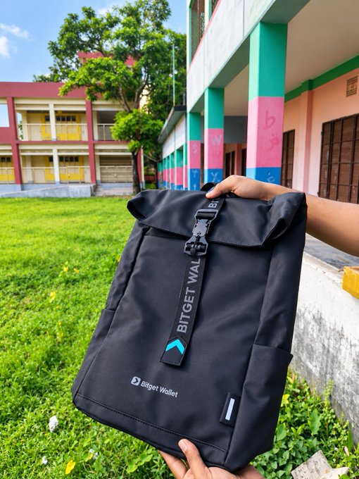 A person holds up a black roll-top backpack with the "Bitget Wallet" logo and a teal chevron design in front of a colorful school building and grassy yard.
