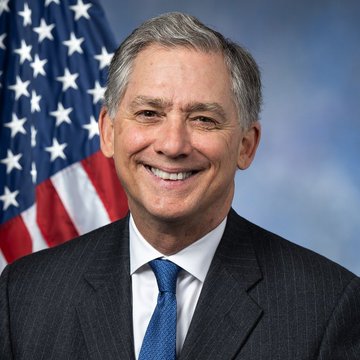 Official portrait of a smiling man in a dark suit and blue tie, standing in front of an American flag.