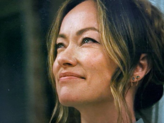 A close-up portrait of a woman with shoulder-length brown hair smiling softly while looking upward against a dark background.