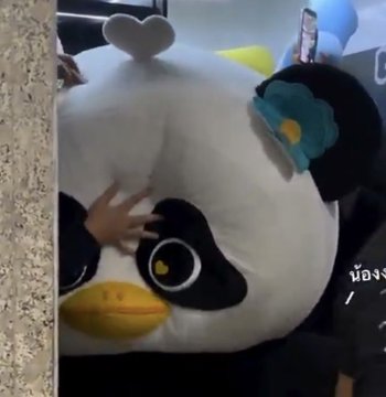 A plush penguin mascot, likely from the K-pop group BTS (known as "Chimmy" or similar BT21 character), is humorously "stuck" against a wall with a hand pressed flat against its face in a comically squished expression, making the heart shape on its forehead appear extra prominent and matching the viral "#LUNAR" meme trend.