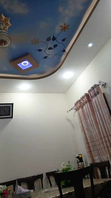 A room interior featuring a decorative blue ceiling with painted clouds and stars, recessed lighting, a chandelier, brown curtains, and dining chairs.