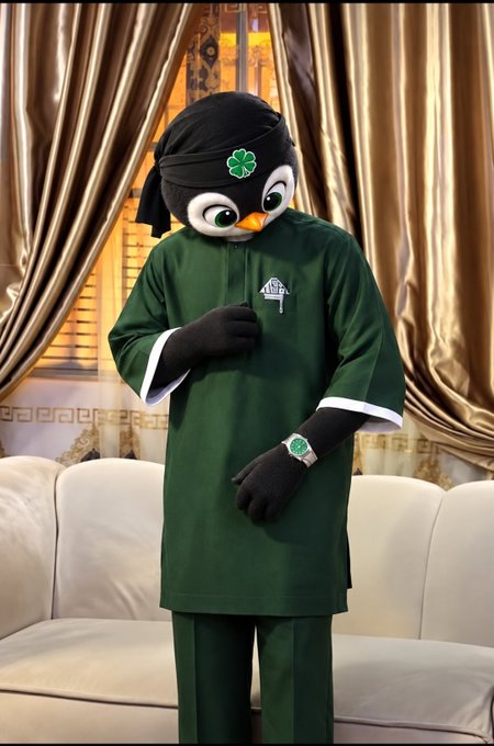 A person dressed in a green mascot costume with a black penguin-like head featuring a four-leaf clover, standing in front of gold curtains on a stage.