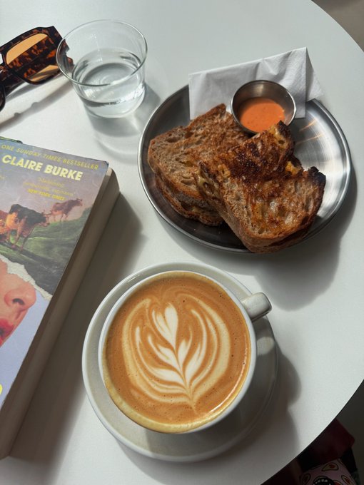 Photo of a cafe table with a latte featuring leaf latte art, grilled cheese/toast with a dipping sauce, a glass of water, and a book by 'CLAIRE BURKE' from the New York Times Bestseller list.