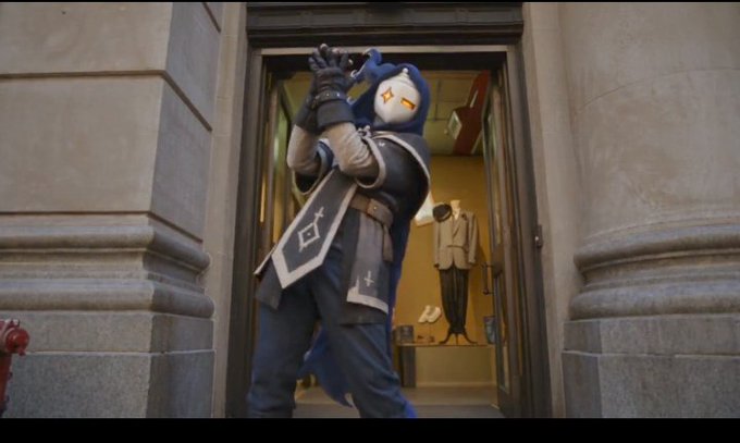A person wearing a stylized blue and gold owl-mask costume and cape stands in a grand doorway, looking through a spyglass.