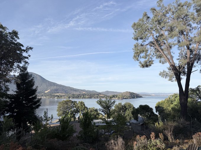 A scenic waterfront view from an elevated position, looking across a calm bay toward forested hills and mountains under a blue sky with wispy clouds.
