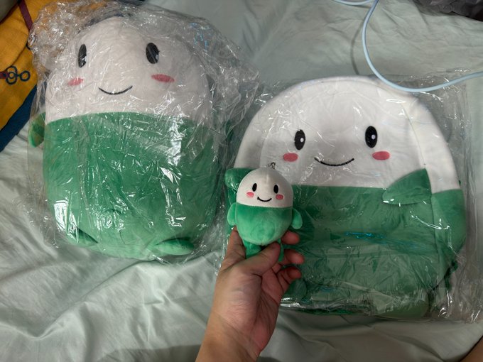Two green and white kawaii-style plush toys sit on a bed in plastic packaging, with a hand holding a matching smaller plushie, perfectly illustrating the "Unwrap me! I can't breathe!" caption as the toys remain sealed in their plastic wrapping.