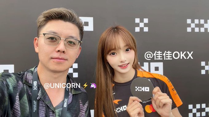 A man with glasses and a lanyard poses for a selfie next to a young woman with long hair at what appears to be an OKX-sponsored esports or gaming event, with the handles "@UNICORN" and "@佳佳OKX" overlaid on the image. The accompanying post contrasts the man's ordinary appearance with the woman's natural beauty, suggesting she looks the same even without beauty filters, and encourages followers to check out her account.