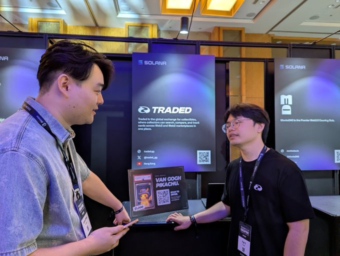 Two men standing in front of banners for Solana-based projects 'TRADED' and 'BlackElk' at a crypto or web3 conference.