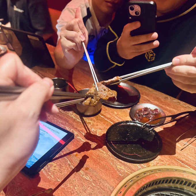 Several people use chopsticks to grab meat from a shared dish at a restaurant table with sauces and a phone visible.