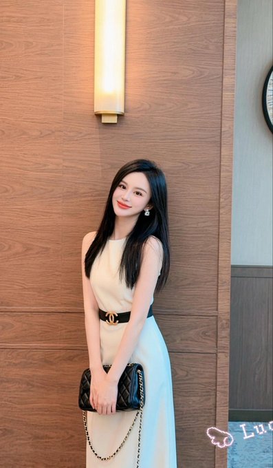 A woman with long black hair poses in a sleeveless cream dress with a Chanel belt, holding a black quilted handbag.