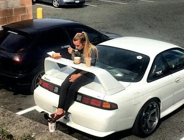 A blonde woman sits on the rear wing/trunk area of a white sports car, eating a meal of fast food and coffee, with a dark hatchback parked next to it in a parking lot.