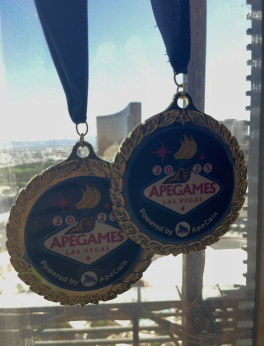 Two medals hang in front of a window overlooking a city skyline, featuring 'APEGAMES LAS VEGAS 2025' and 'Powered by ApeCoin' text with a casino chip design.