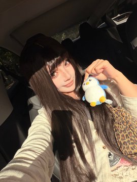 A selfie of a young woman with long dark hair wearing a leopard print top, sitting in a car and holding a small plush toy near her face.