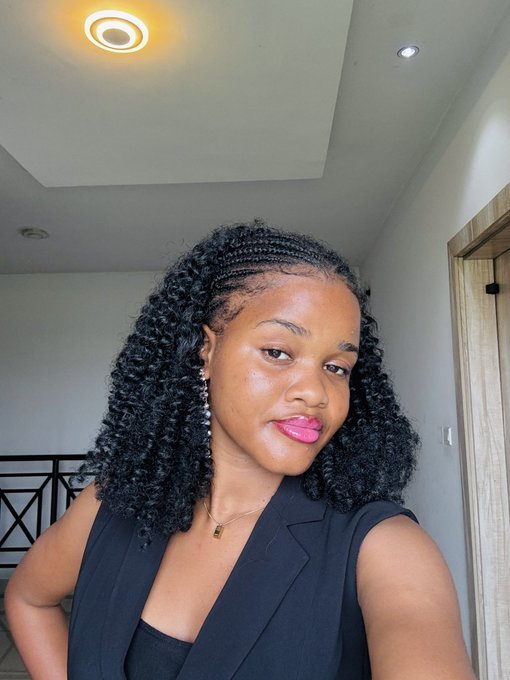 A woman with braided and curly hair, wearing a sleeveless black blazer and bright pink lipstick, poses for a selfie indoors as a cheerful "Gm." (good morning) greeting.