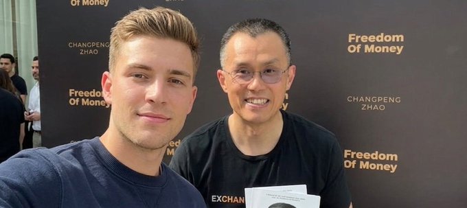 Two men pose for a photo in front of a "Changpeng Zhao: Freedom of Money" event backdrop, with the older man on the right identified as CZ (Changpeng Zhao), founder of Binance, wearing a black EXCHANGE shirt and holding a book, while a younger man on the left smiles for the selfie—capturing the "GM CT" (good morning Crypto Twitter) greeting posted by the younger attendee after meeting the crypto industry figure.