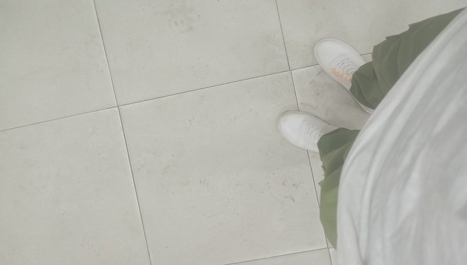 A downward-facing photo showing a person's feet in white sneakers and green pants standing on light-colored tiled flooring.