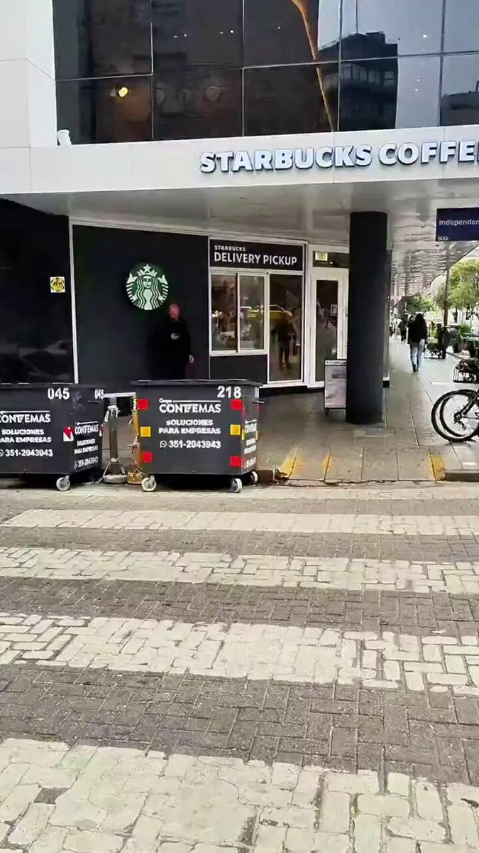 The image shows a Starbucks coffee shop with its name displayed above the entrance, indicating that it's open for business. The exterior of the building features a black and white striped sidewalk, which contrasts with the modern design of the Starbucks store. A person is standing on the sidewalk in front of the Starbucks, possibly waiting to enter or just passing by. There are also two suitcases near the entrance, suggesting that someone might be traveling or has recently arrived at the location.