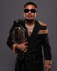 Joshua Van, wearing sunglasses and a black blazer with colorful trim, proudly displays a UFC championship belt in a promotional photo for UFC 331. The image ties to the post's question about whether he will defend his title against Alexandre Pantoja.
