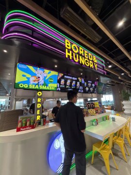 A brightly lit "Bored Hungry" food counter/restaurant with neon signage where a person in a black hoodie stands at the ordering area.
