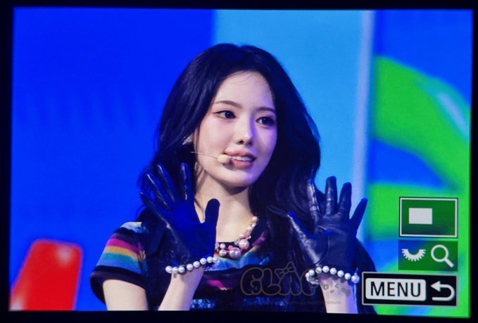A woman with dark hair wearing black gloves performs on stage, shown on a TV screen display with a 'MENU' button overlay in the corner.