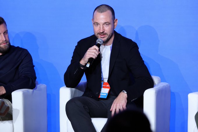 A bald man in a black suit speaks into a microphone while seated on a white chair on stage with a blue backdrop, wearing a lanyard with a badge.