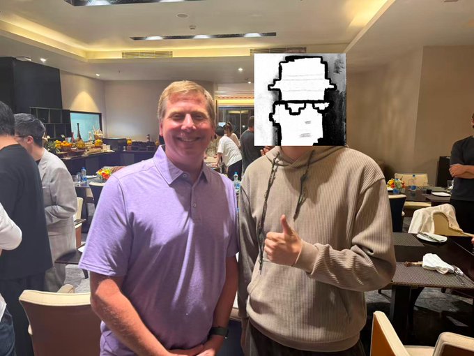 Two men pose together at an indoor event, with a smiling man in a purple shirt standing next to a person in a grey hoodie whose face is covered by a pixelated graphic.
