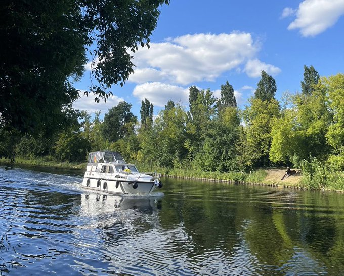 A white motorboat cruising along a calm river lined with lush green trees under a blue sky with scattered white clouds.
